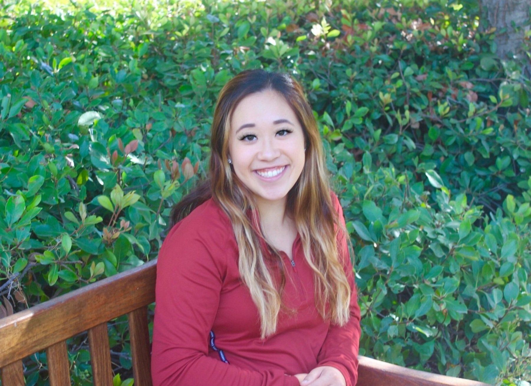 Ashley Tai, smiling, seated on a wooden bench in front of green hedges.